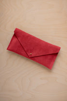 A scarlet red suede envelope clutch wallet or bag with a small gold button clasp, resting on a light beige wood grain background.