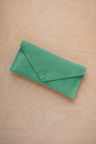 A seafoam turquoise green coloured suede envelope clutch wallet or bag with a small gold button clasp, resting on a light beige wood grain background.
