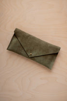A soft olive green coloured suede envelope clutch wallet or bag with a small gold button clasp, resting on a light beige wood grain background.