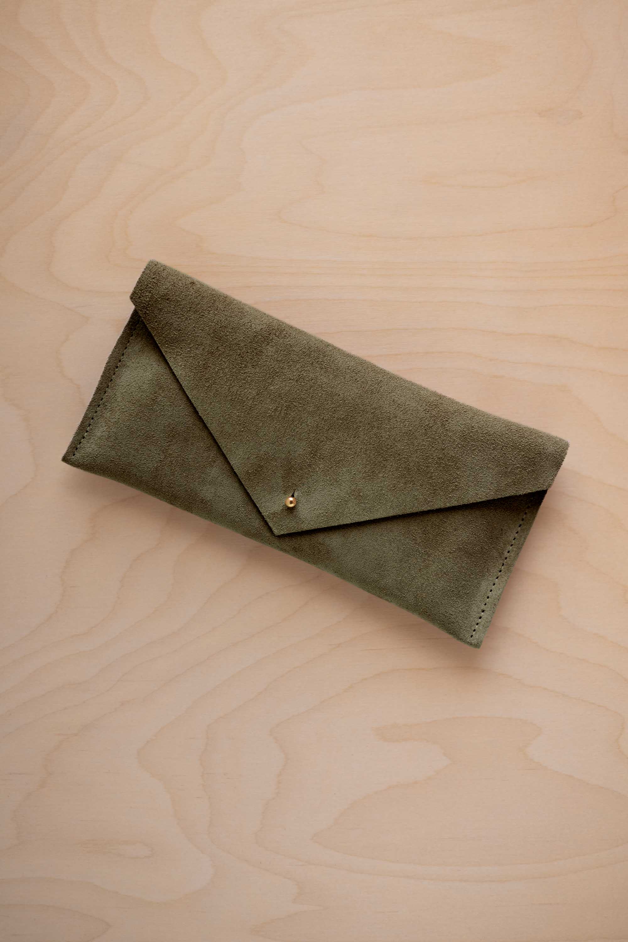 A soft olive green coloured suede envelope clutch wallet or bag with a small gold button clasp, resting on a light beige wood grain background.