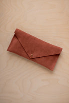 A terracotta coloured envelope clutch wallet or bag with a small gold button clasp, resting on a light beige wood grain background.
