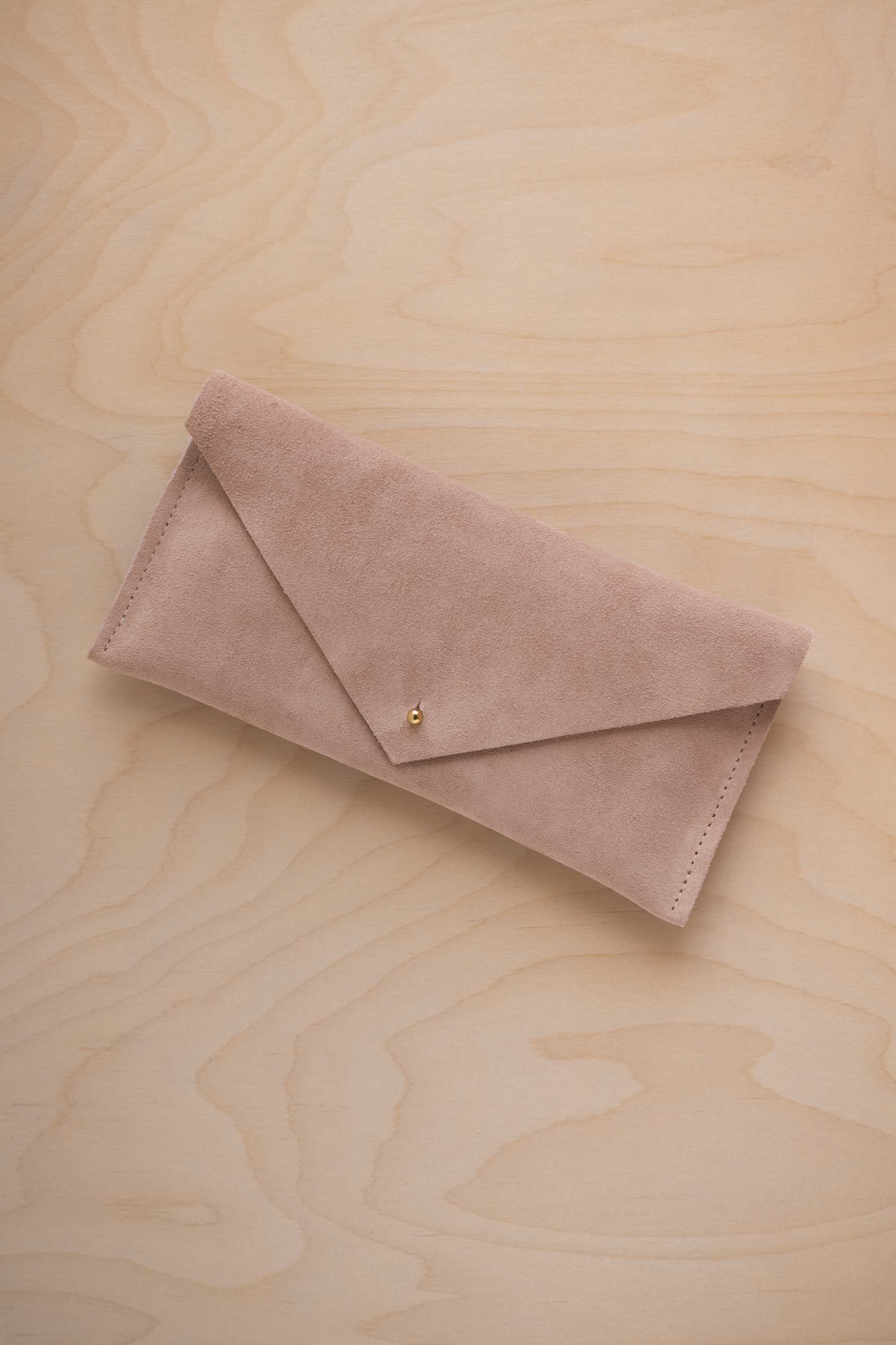 A beige suede envelope clutch wallet or bag with a small gold button clasp, resting on a light beige wood grain background.