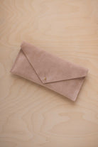 A beige suede envelope clutch wallet or bag with a small gold button clasp, resting on a light beige wood grain background.