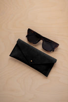 A pair of dark tortoiseshell sunglasses sit next to a black suede envelope style eye glasses case with a small gold button, set on a light wood grain background.