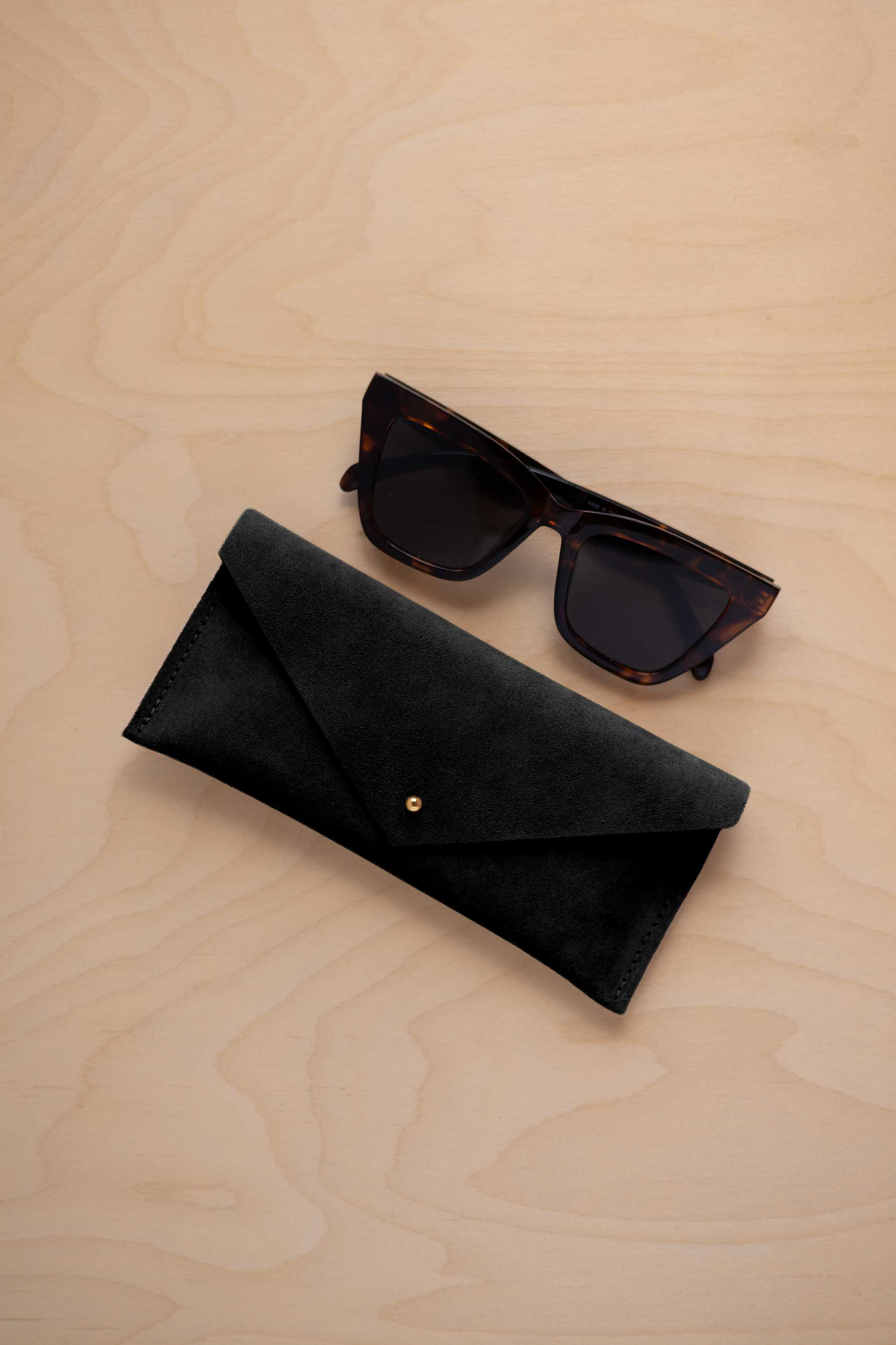 A pair of dark tortoiseshell sunglasses sit next to a black suede envelope style eye glasses case with a small gold button, set on a light wood grain background.