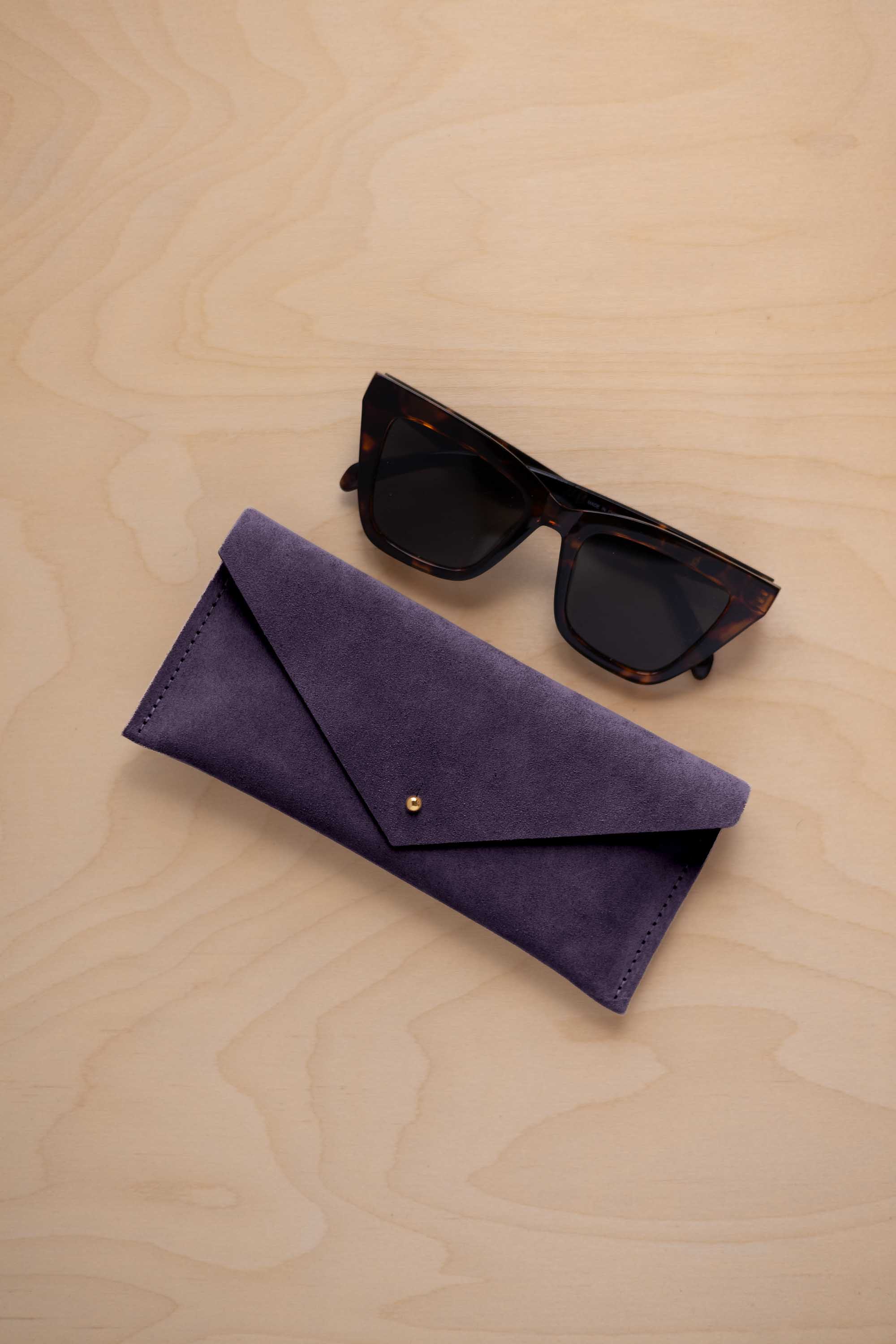 A pair of dark tortoiseshell sunglasses sit next to a blackcurrent dark purple suede envelope style eye glasses case with a small gold button, set on a light wood grain background.