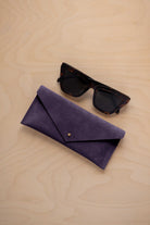 A pair of dark tortoiseshell sunglasses sit next to a blackcurrent dark purple suede envelope style eye glasses case with a small gold button, set on a light wood grain background.