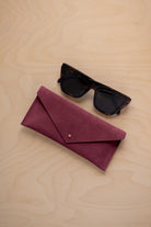 A pair of dark tortoiseshell sunglasses sit next to a burgundy suede envelope style eye glasses case with a small gold button, set on a light wood grain background.