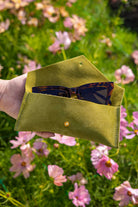 A hand holds an open chartreuse green suede sunglasses case with a small gold button, containing tortoiseshell sunglasses, set against a blurred background of pink flowers and green grass.