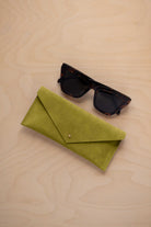 A pair of dark tortoiseshell sunglasses sit next to a chartreuse lime green suede envelope style eye glasses case with a small gold button, set on a light wood grain background.