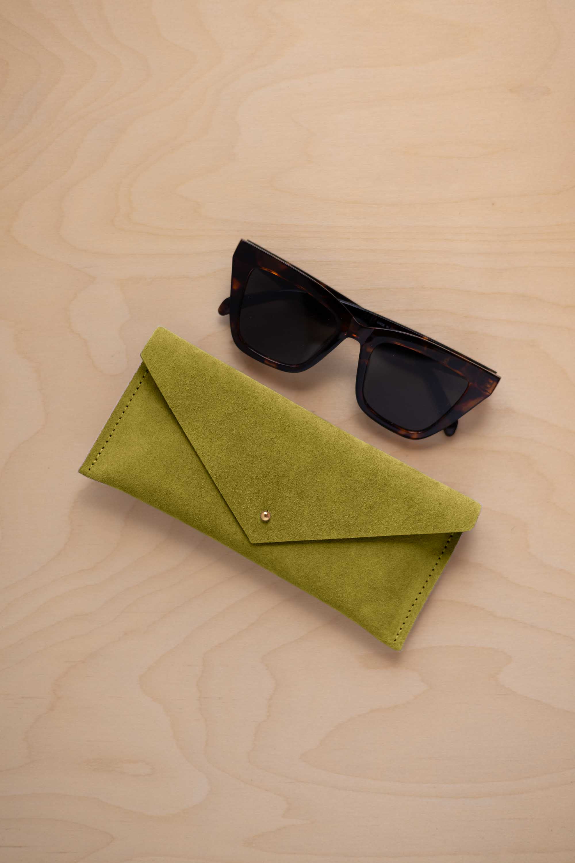 A pair of dark tortoiseshell sunglasses sit next to a chartreuse lime green suede envelope style eye glasses case with a small gold button, set on a light wood grain background.