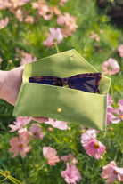 A hand holds an open chartreuse green suede sunglasses case with a small gold button, containing tortoiseshell sunglasses, set against a blurred background of pink flowers and green grass.