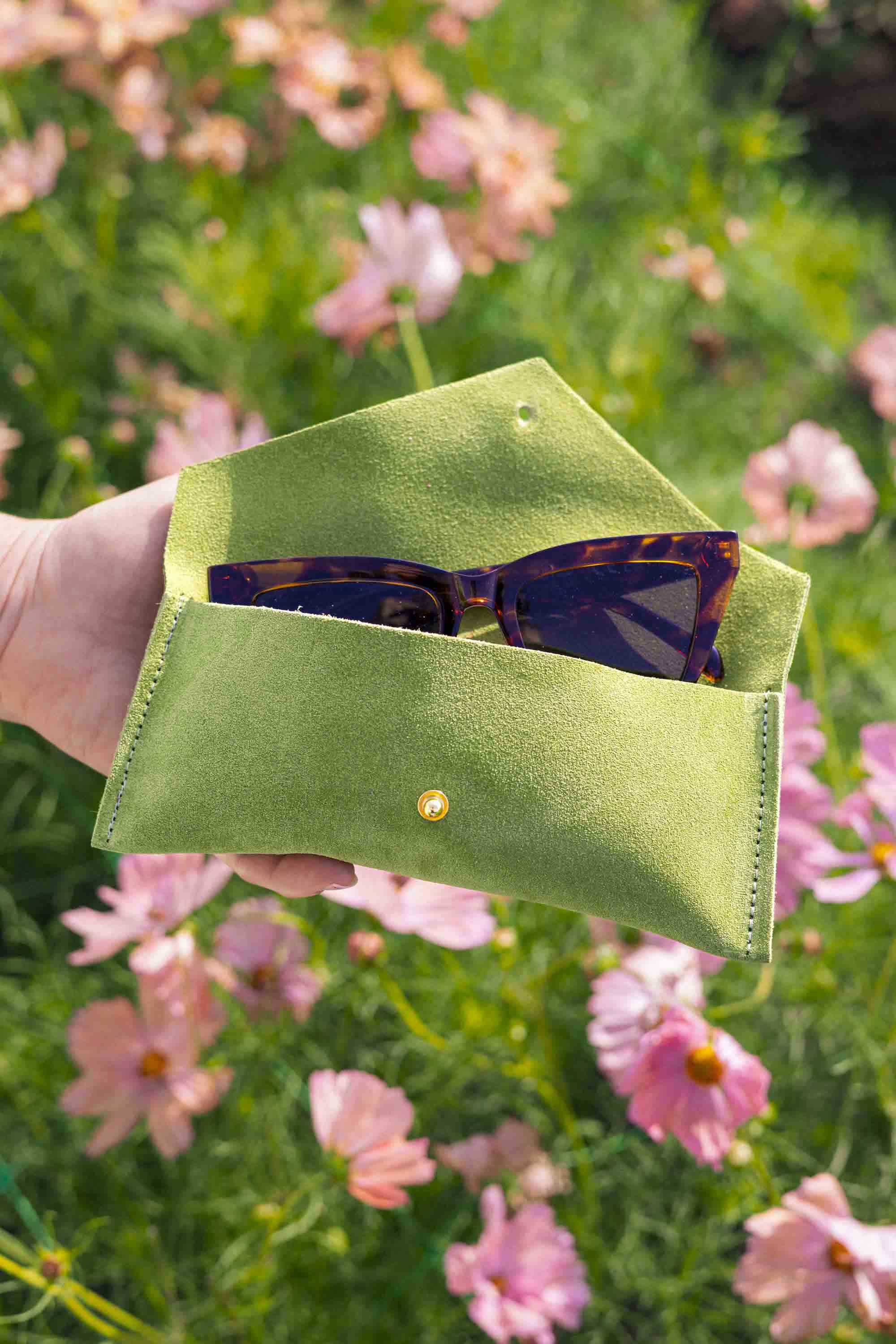 A hand holds an open chartreuse green suede sunglasses case with a small gold button, containing tortoiseshell sunglasses, set against a blurred background of pink flowers and green grass.