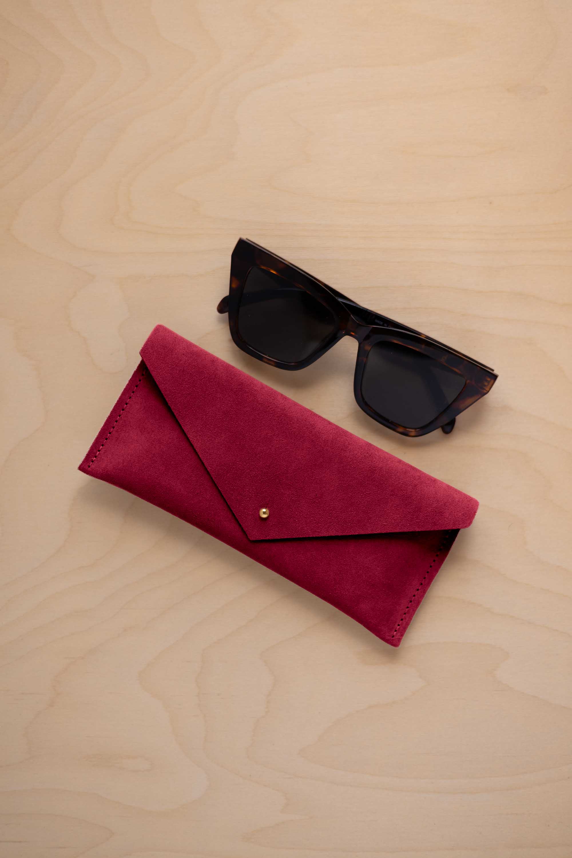 A pair of dark tortoiseshell sunglasses sit next to a crimson red suede envelope style eye glasses case with a small gold button, set on a light wood grain background.