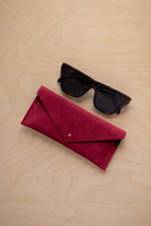 A pair of dark tortoiseshell sunglasses sit next to a crimson red suede envelope style eye glasses case with a small gold button, set on a light wood grain background.