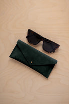 A pair of dark tortoiseshell sunglasses sit next to a deep forest green suede envelope style eye glasses case with a small gold button, set on a light wood grain background.