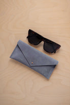 A pair of dark tortoiseshell sunglasses sit next to a dusky blue suede envelope style eye glasses case with a small gold button, set on a light wood grain background.