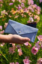 A light blue suede envelope-style eye glasses case with a small gold button, held in a person's hand against a blurred background of pink and purple flowers in a green field.