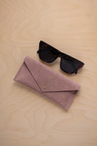 A pair of dark tortoiseshell sunglasses sit next to a dusty mauve grey suede envelope style eye glasses case with a small gold button, set on a light wood grain background.