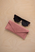 A pair of dark tortoiseshell sunglasses sit next to a dusty rose pink suede envelope style eye glasses case with a small gold button, set on a light wood grain background.