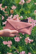 A hand holding a dusty pink envelope-style suede eyeglasses case with a small gold button, set against a blurred background of pink flowers and green foliage.