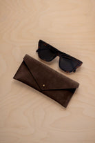 A pair of dark tortoiseshell sunglasses sit next to an espresso brown coloured suede envelope style eye glasses case with a small gold button, set on a light wood grain background.