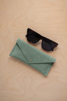 A pair of dark tortoiseshell sunglasses sit next to a eucalyptus sage green coloured suede envelope style eye glasses case with a small gold button, set on a light wood grain background.