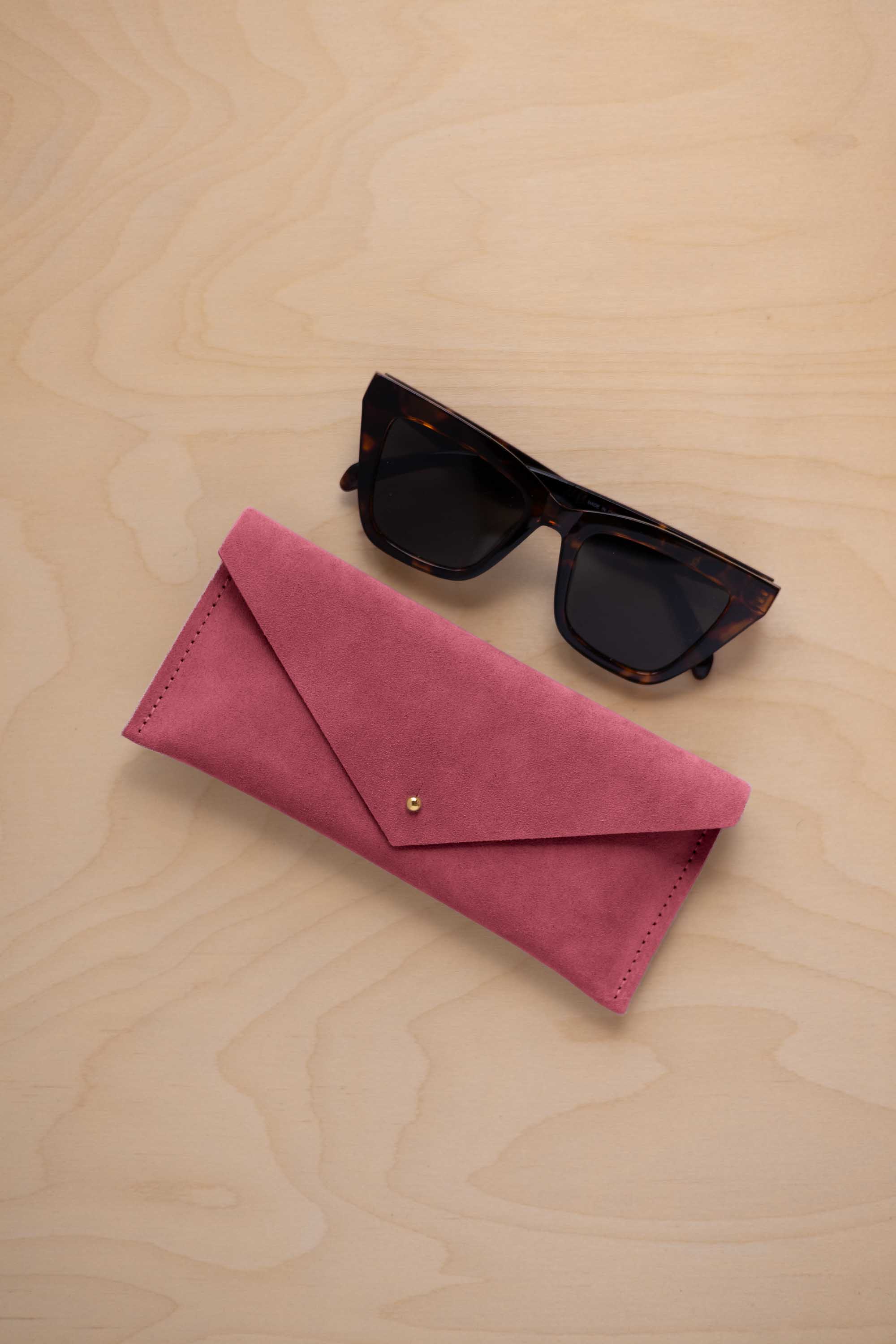 A pair of dark tortoiseshell sunglasses sit next to a faded rose pink suede envelope style eye glasses case with a small gold button, set on a light wood grain background.