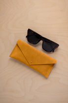 A pair of dark tortoiseshell sunglasses sit next to a mustard yellow suede envelope style eye glasses case with a small gold button, set on a light wood grain background.