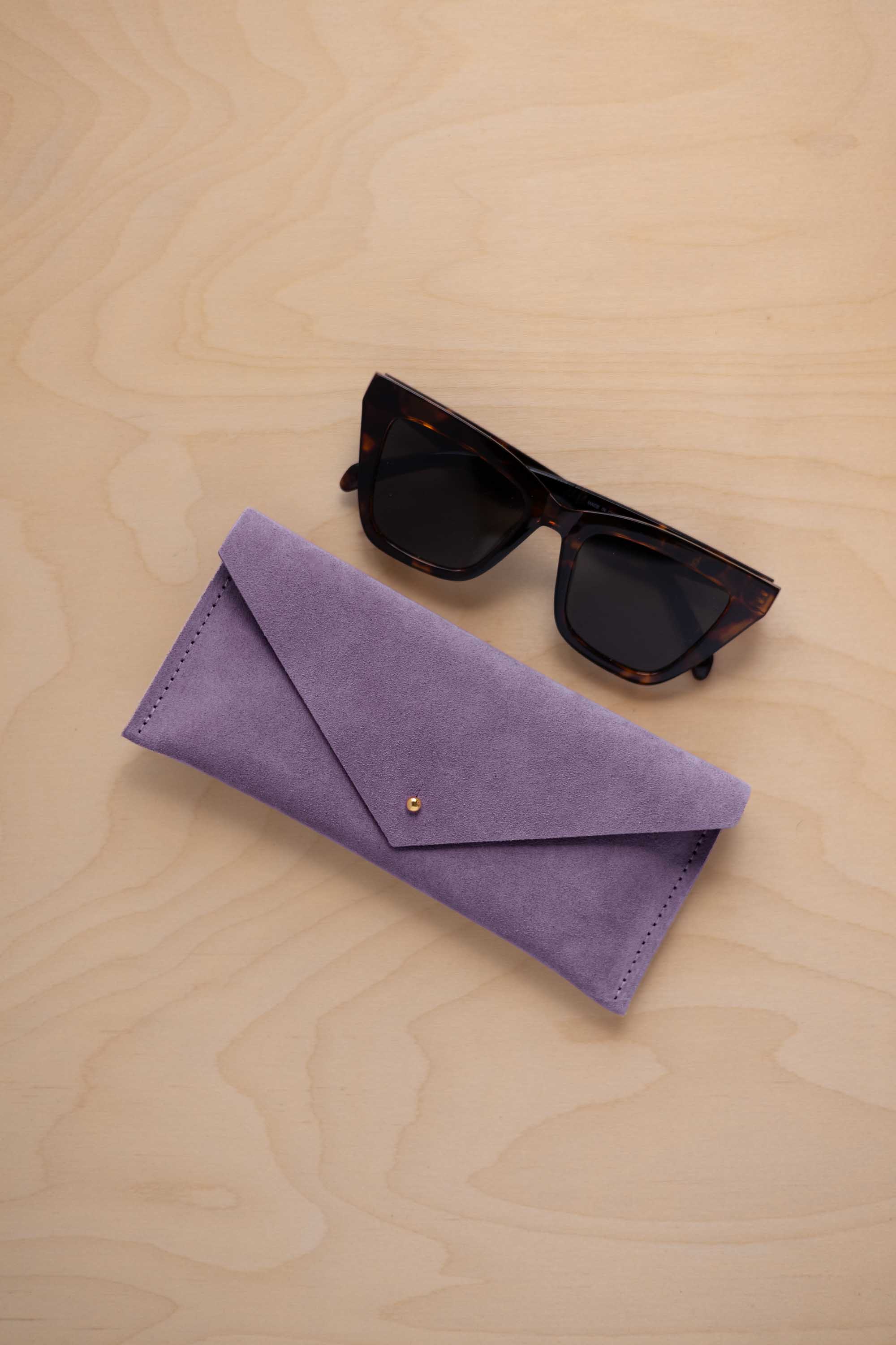 A pair of dark tortoiseshell sunglasses sit next to a lavender suede envelope style eye glasses case with a small gold button, set on a light wood grain background.