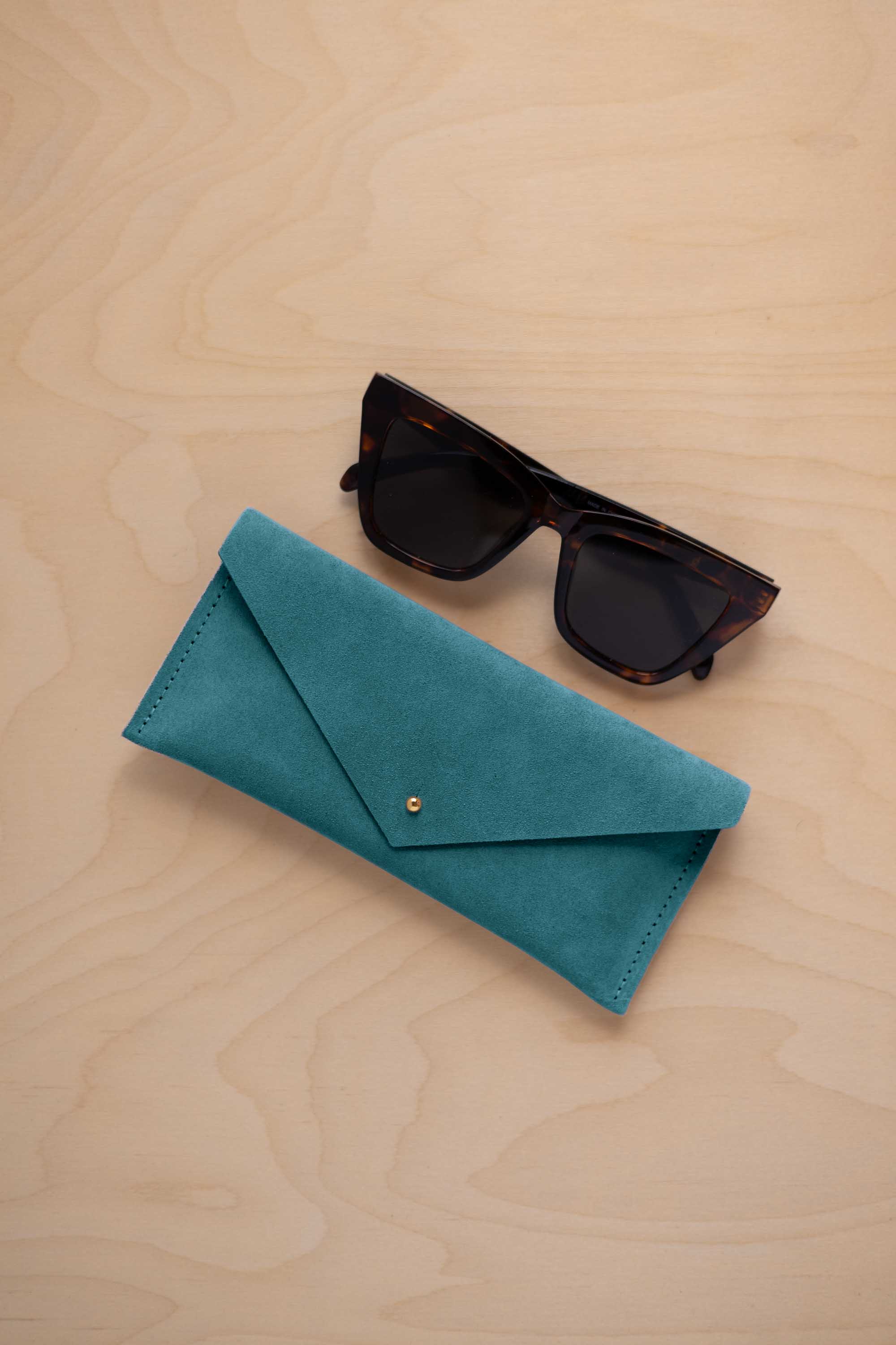 A pair of dark tortoiseshell sunglasses sit next to a marine teal coloured suede envelope style eye glasses case with a small gold button, set on a light wood grain background.