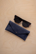 A pair of dark tortoiseshell sunglasses sit next to a midnight navy coloured suede envelope style eye glasses case with a small gold button, set on a light wood grain background.