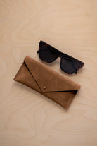 A pair of dark tortoiseshell sunglasses sit next to a mocha brown suede envelope style eye glasses case with a small gold button, set on a light wood grain background.