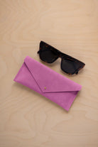 A pair of dark tortoiseshell sunglasses sit next to a peony pink suede envelope style eye glasses case with a small gold button, set on a light wood grain background.