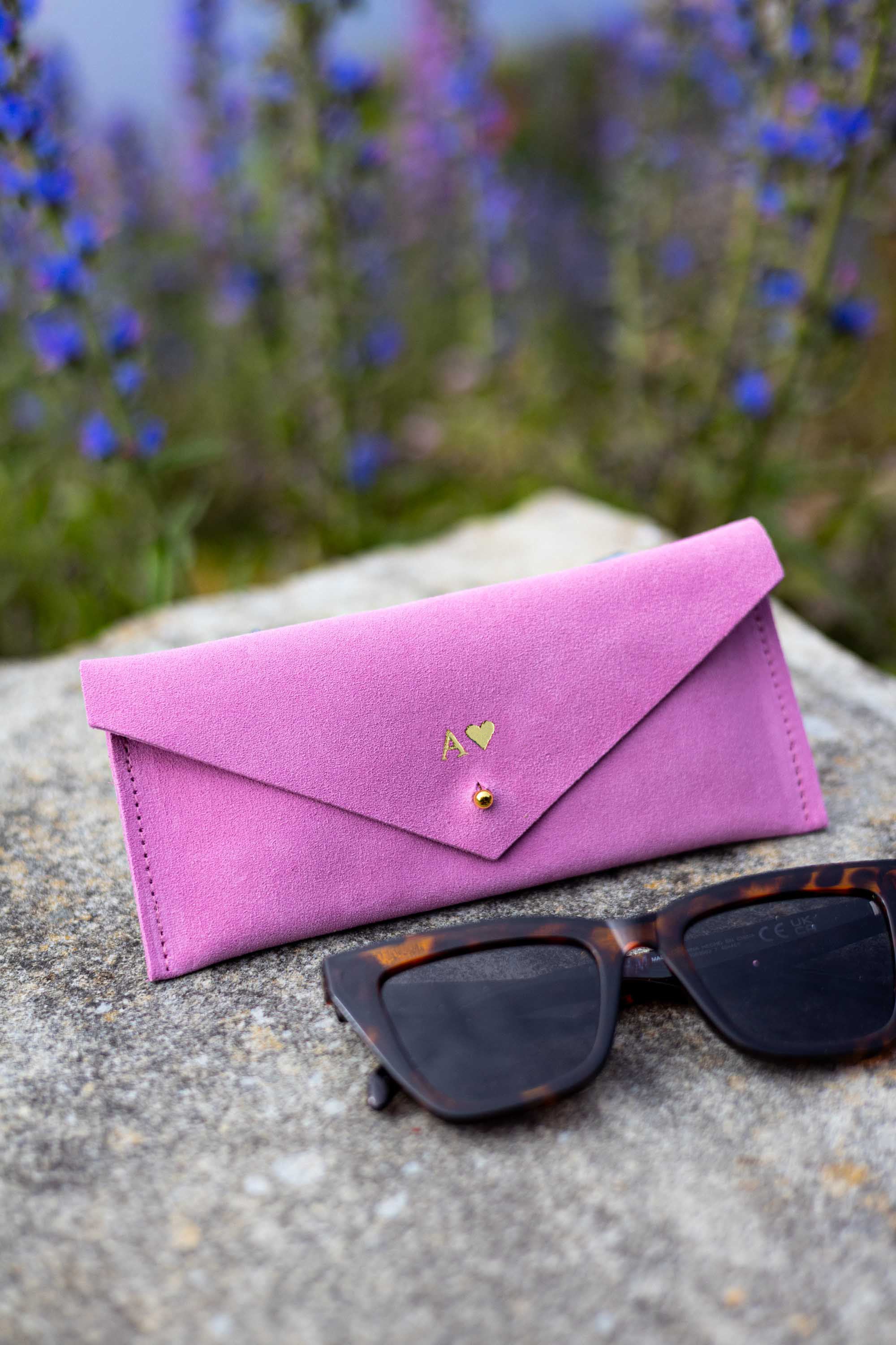 A pink suede envelope-style sunglasses case personalised with a small gold heart logo and the initial A, placed on a stone surface outdoors with tortoiseshell sunglasses in front and blurred blue flowers in the background.