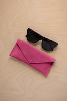 A pair of dark tortoiseshell sunglasses sit next to a raspberry pink suede envelope style eye glasses case with a small gold button, set on a light wood grain background.