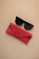 A pair of dark tortoiseshell sunglasses sit next to a scarlet red suede envelope style eye glasses case with a small gold button, set on a light wood grain background.