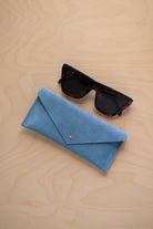 A pair of dark tortoiseshell sunglasses sit next to a sky blue suede envelope style eye glasses case with a small gold button, set on a light wood grain background.