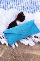 A bright blue envelope-style sunglasses case with a small gold button, styled on a white fringed blanket over sand with tortoiseshell sunglasses resting on top.