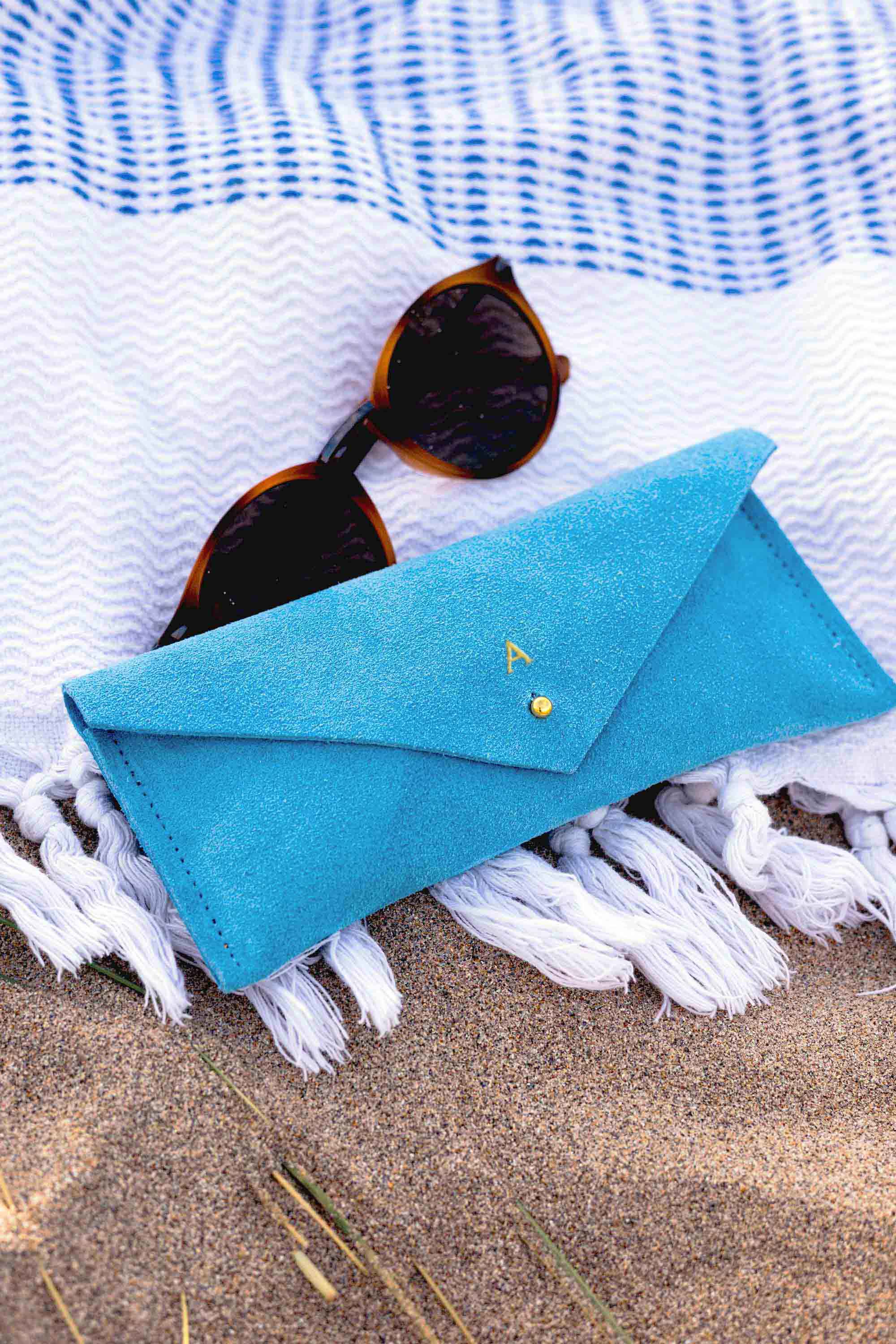 A bright blue envelope-style sunglasses case with a small gold button, styled on a white fringed blanket over sand with tortoiseshell sunglasses resting on top.