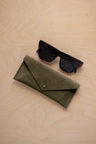 A pair of dark tortoiseshell sunglasses sit next to a soft olive green coloured suede envelope style eye glasses case with a small gold button, set on a light wood grain background.