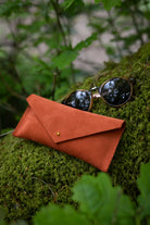 A terracotta orange suede envelope-style sunglasses case with a small gold stud closure, shown outdoors on mossy green foliage with a pair of sunglasses resting on top.