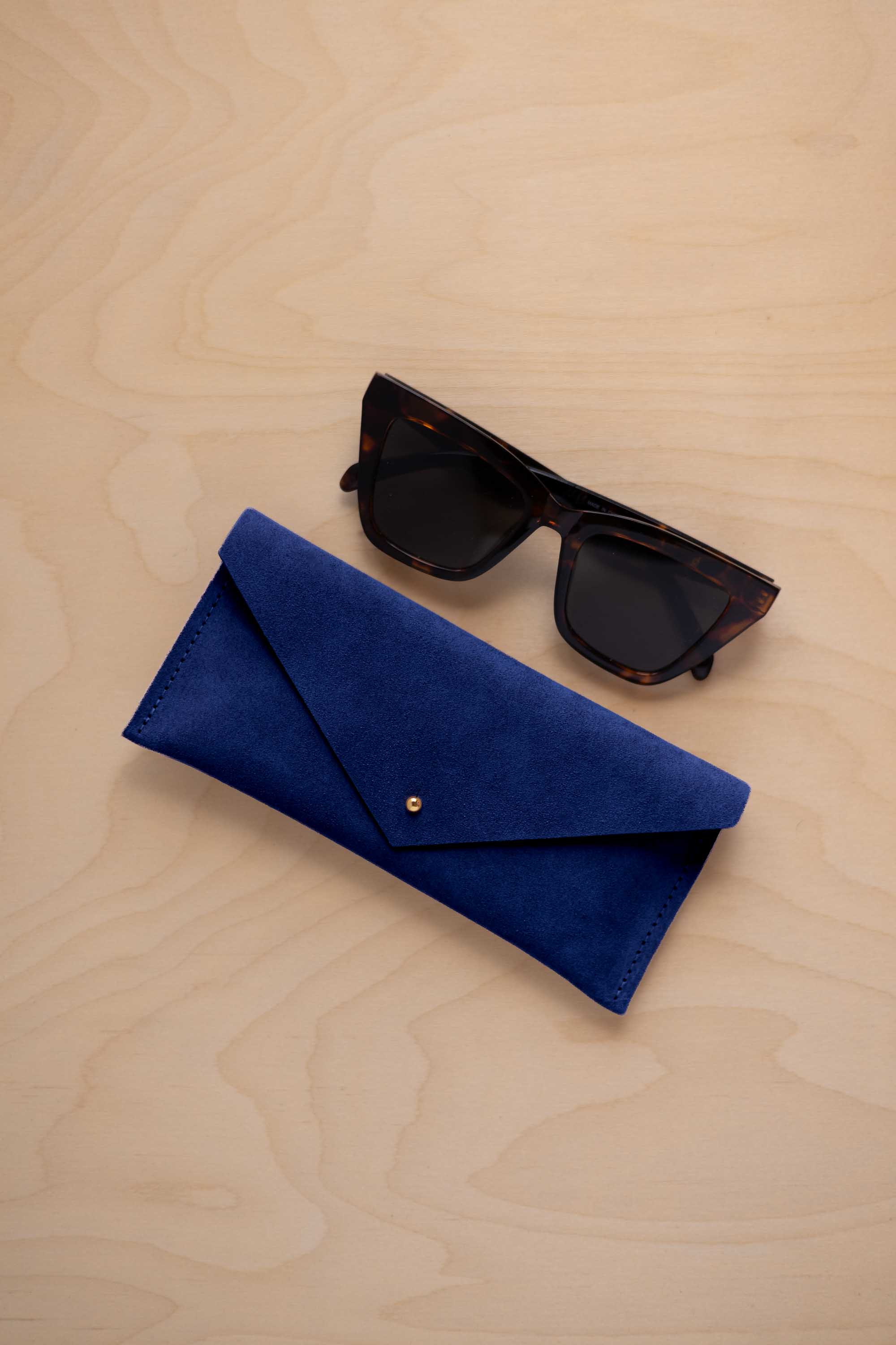 A pair of dark tortoiseshell sunglasses sit next to a ultramarine blue suede envelope style eye glasses case with a small gold button, set on a light wood grain background.