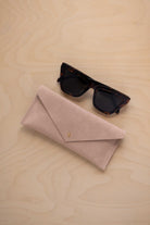 A pair of dark tortoiseshell sunglasses sit next to a warm beige suede envelope style eye glasses case with a small gold button, set on a light wood grain background.