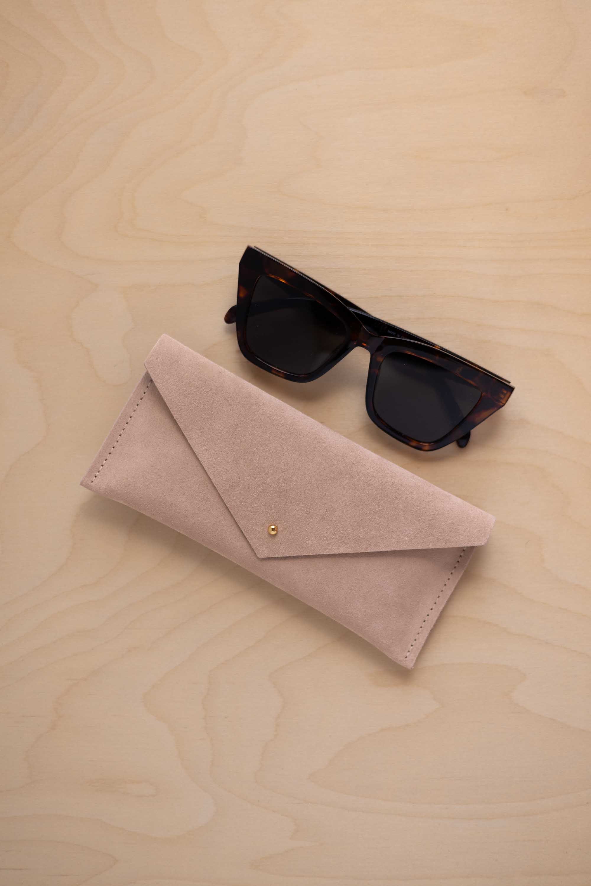 A pair of dark tortoiseshell sunglasses sit next to a warm beige suede envelope style eye glasses case with a small gold button, set on a light wood grain background.