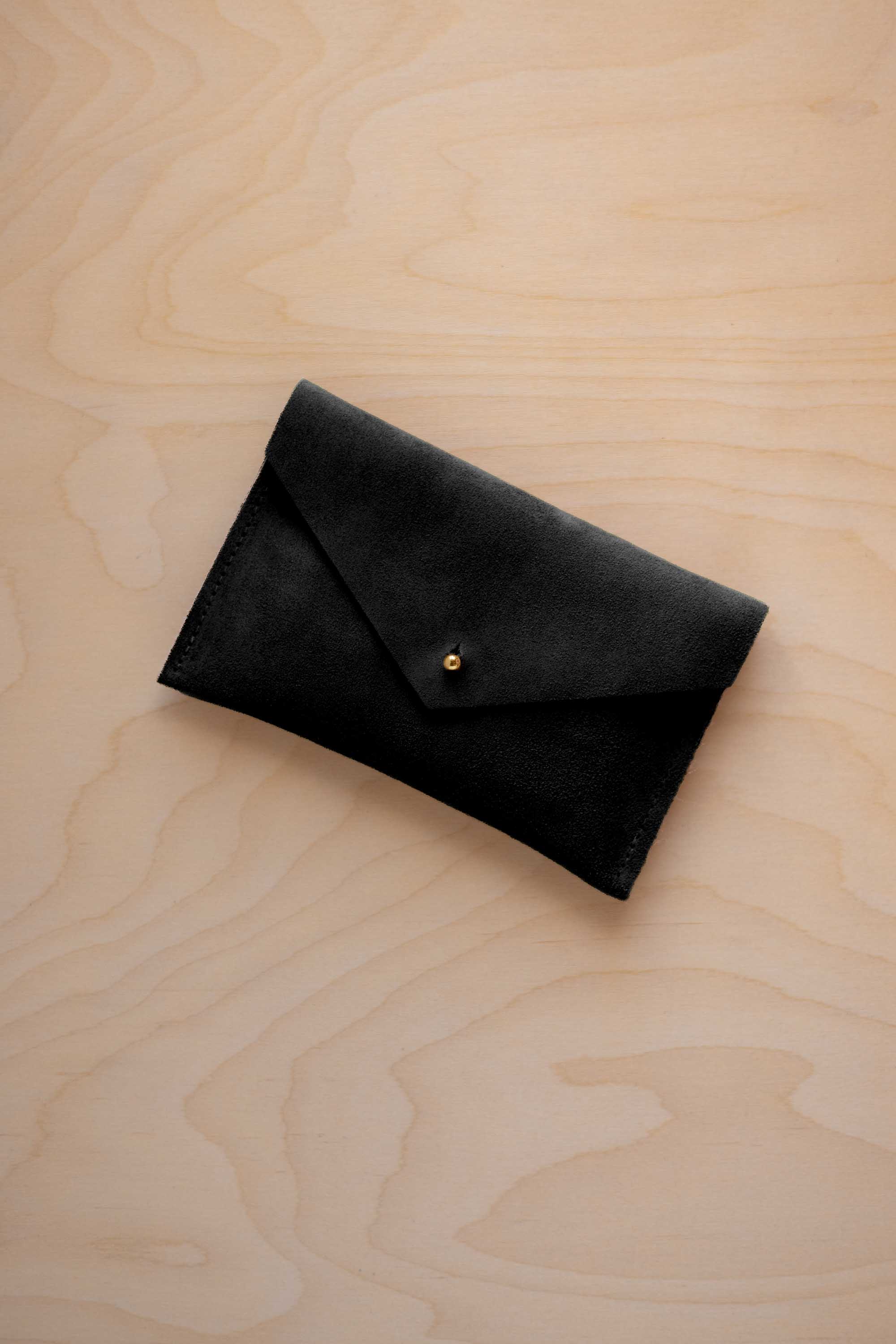 A small black coloured suede envelope-style pouch with a gold button, resting on a light wood grain surface.