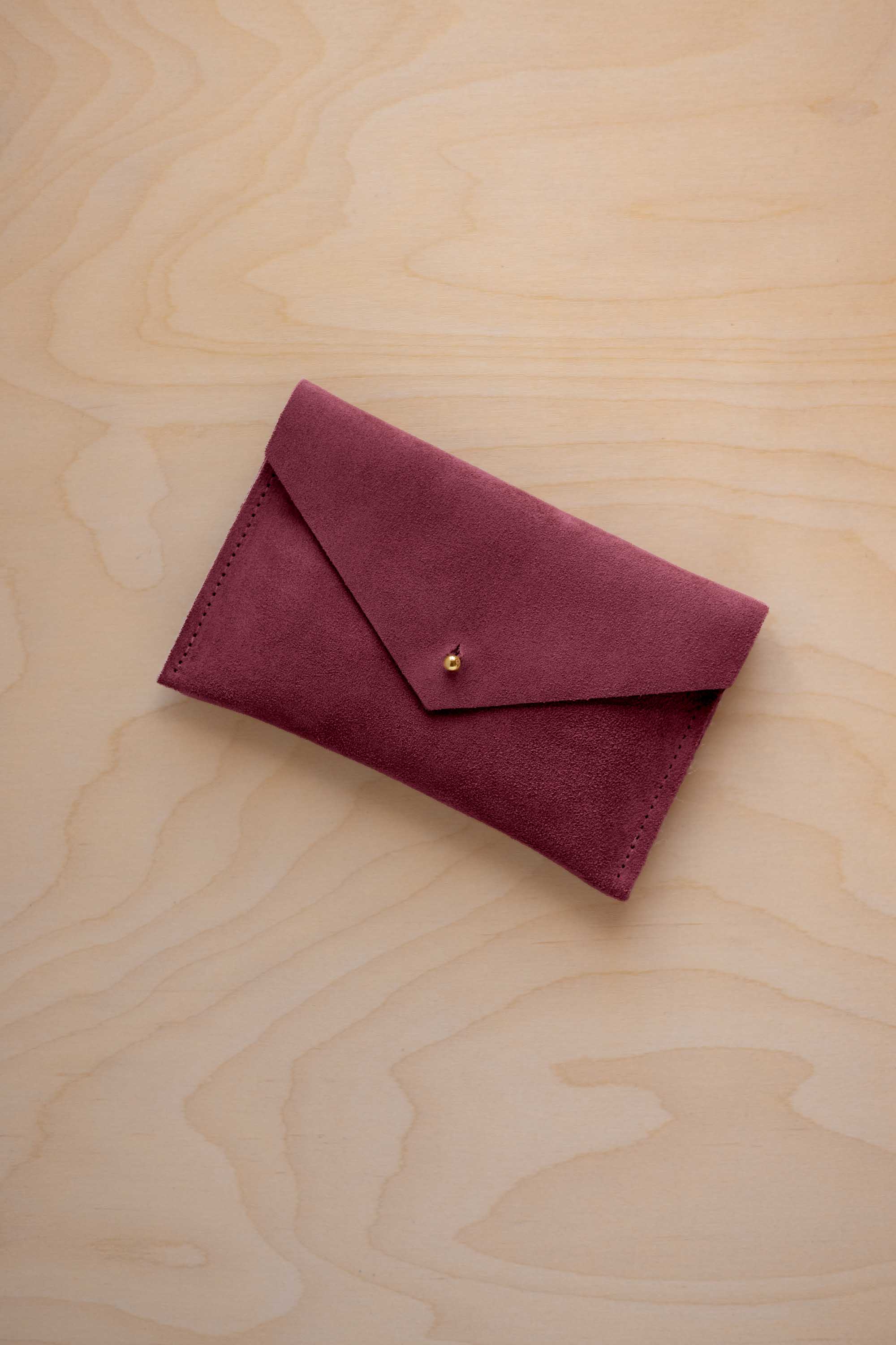 A small burgundy coloured suede envelope-style pouch with a gold button, resting on a light wood grain surface.