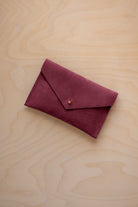 A small burgundy coloured suede envelope-style pouch with a gold button, resting on a light wood grain surface.