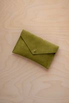 A small chartreuse green coloured suede envelope-style pouch with a gold button, resting on a light wood grain surface.