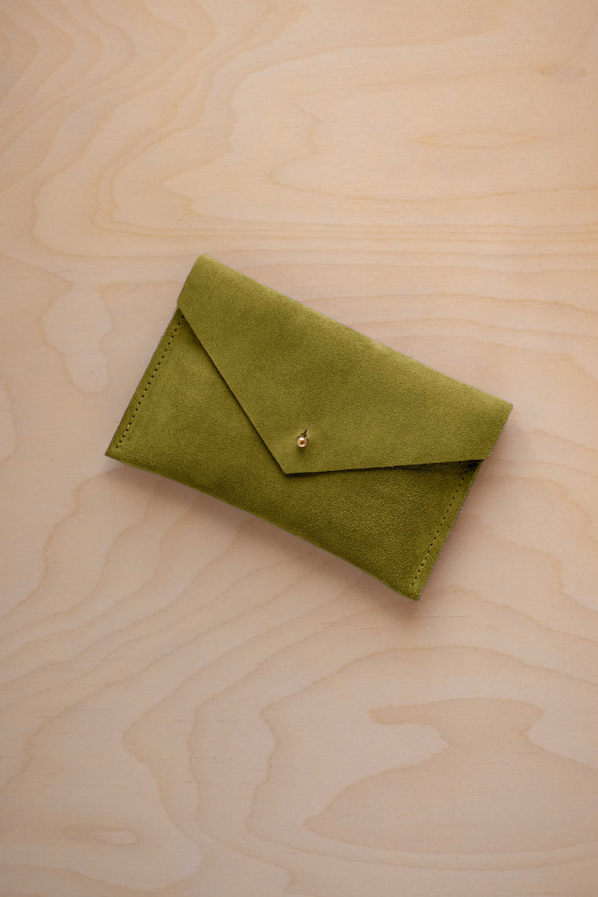 A small chartreuse green coloured suede envelope-style pouch with a gold button, resting on a light wood grain surface.